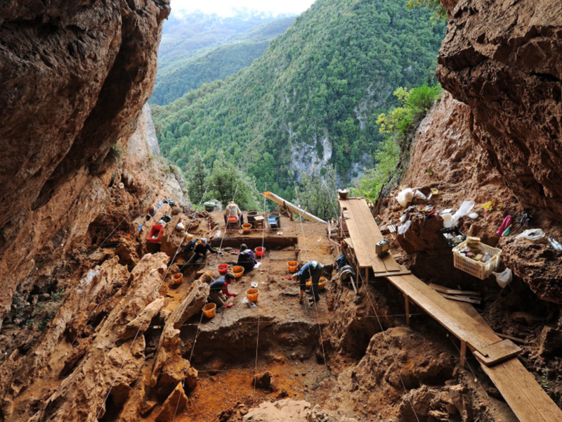 L'immagine mostra un sito archeologico preistorico situato all'interno di una grotta. Nella parte anteriore, l'ingresso della grotta offre una vista panoramica su una valle verdeggiante e montuosa, indicando che la grotta si trova in una zona elevata e isolata. Dettagli dell'immagine: Sito di scavo: Nella parte centrale e inferiore dell'immagine, si vedono diversi archeologi al lavoro, intenti a scavare e documentare il sito. Il terreno è suddiviso in griglie, probabilmente per facilitare la catalogazione dei reperti e la documentazione stratigrafica. Gli archeologi sono circondati da attrezzature di scavo, tra cui secchi, cassette e strumenti per scavare. Strutture di supporto: Sulla destra, c'è una passerella di legno sopraelevata che permette agli archeologi di muoversi sopra l'area di scavo senza disturbare il terreno sottostante. Ci sono tavole e contenitori organizzati lungo i lati della grotta, usati probabilmente per raccogliere e analizzare i reperti. Ambiente naturale: La grotta ha un'ampia apertura che si affaccia su un paesaggio naturale lussureggiante, con colline ricoperte di alberi e vegetazione. L'ambiente sembra isolato e tranquillo, ideale per un sito archeologico che richiede concentrazione e cura. Questa immagine evidenzia l'importanza delle grotte come luoghi di ricerca archeologica, dove gli studiosi possono scoprire e studiare i resti delle antiche occupazioni umane e comprendere meglio la vita preistorica.